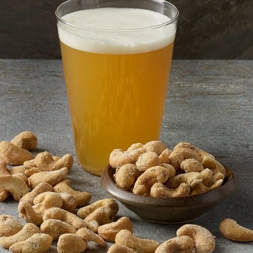 Glass of beer with a bowl of cashews on a table.